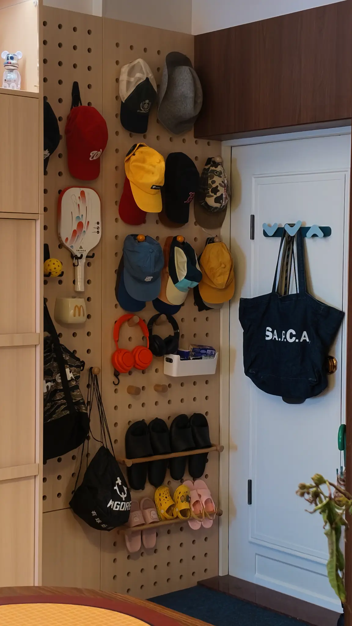 Perforated timber entry wall holding caps, bags, headphones and footwear beside a white door.