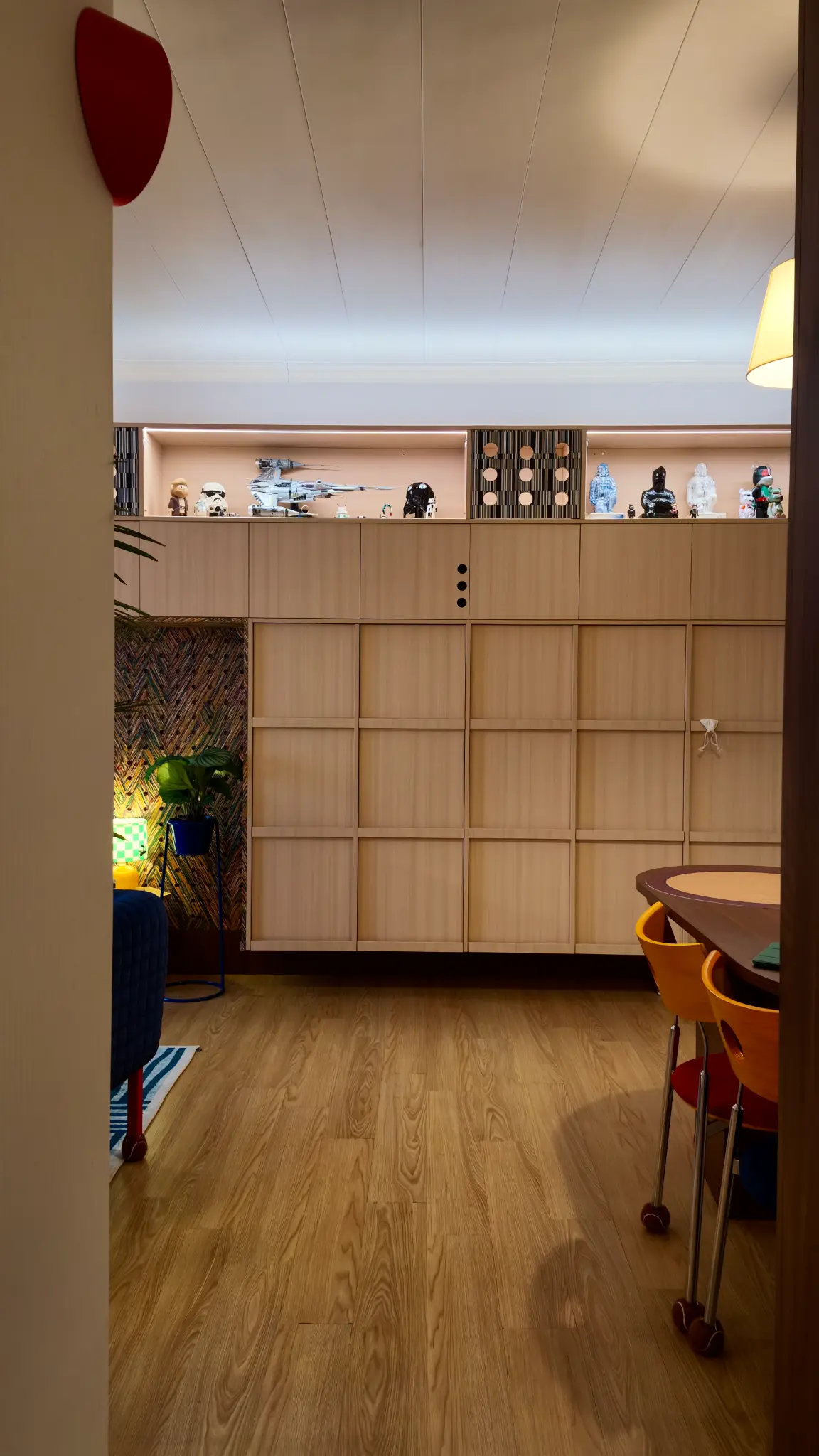 View into a room with pale timber storage, shelves, a patterned wall and dining seating.