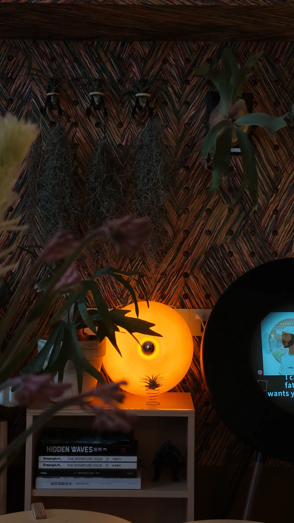 A glowing round orange lamp against a richly patterned wall with plants and books.