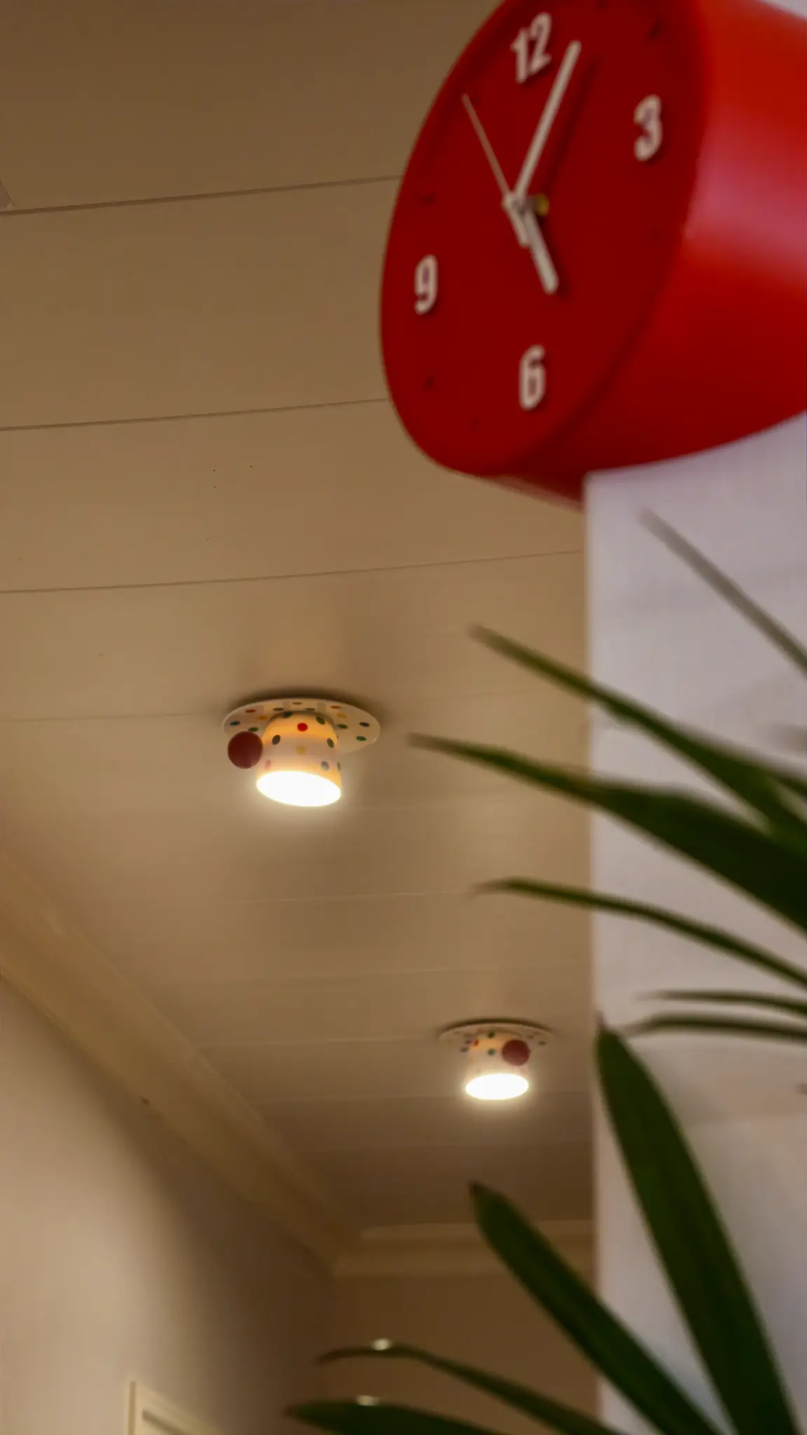 Red wall clock and decorative ceiling lights viewed through plant leaves.