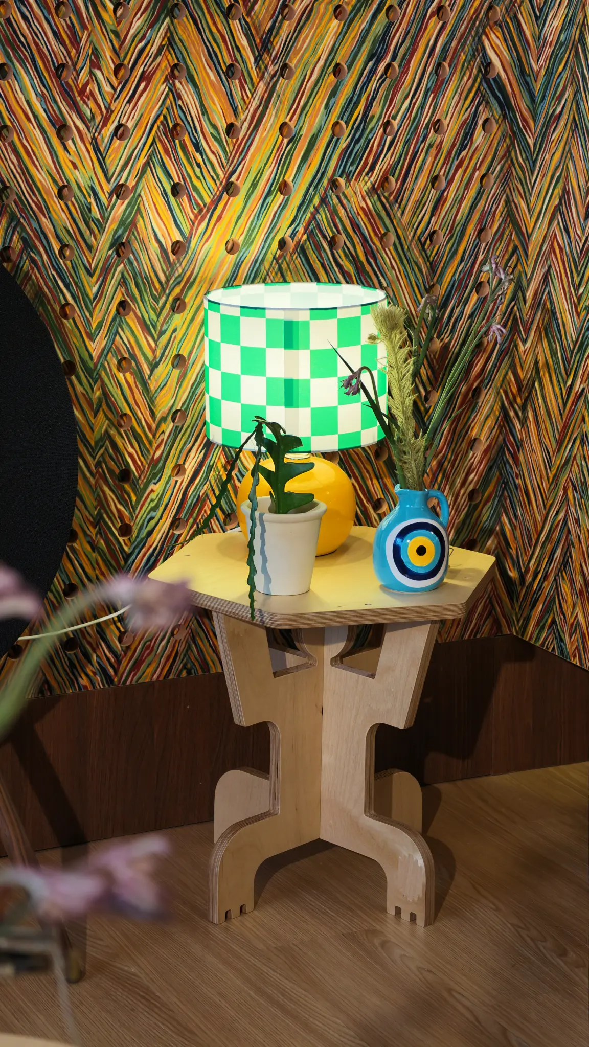 Green checkerboard lamp, blue vase and timber side table before a colourful textured wall.